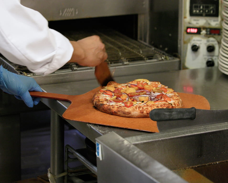 Chef in gloves sliding a fresh baked pizza on a wooden peel at a stainless commercial pizza station